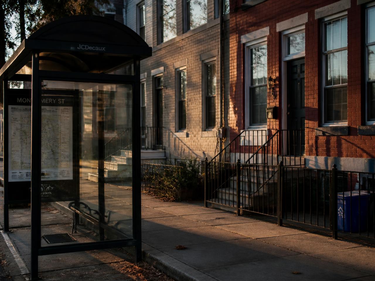 Rowhouses and a bus shelter on a neighborhood sidewalk in late light